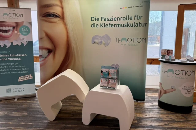 Messestand von TJ-Motion mit Roll-ups, Produktdisplay und Sitzmöbeln zur Präsentation der Faszienrolle für die Kiefermuskulatur.