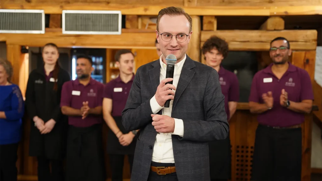 Ein Mann spricht mit einem Mikrofon zu den Gästen, im Hintergrund steht ein Team in einheitlicher Kleidung.