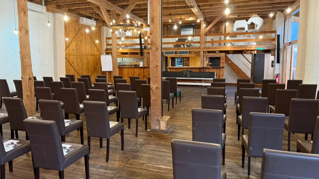 Bestuhlter Veranstaltungsraum mit Reihen aus Stühlen in einer rustikalen Halle, vorbereitet für einen Vortrag oder eine Präsentation.