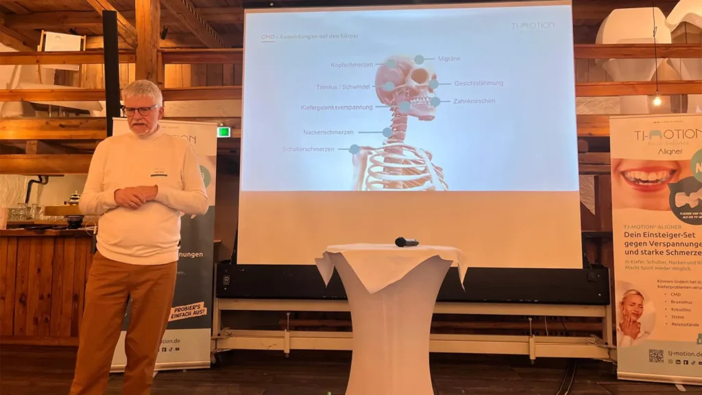 Jürgen Meier steht in einem Holzsaal vor einer Leinwand mit einer Grafik zu Kopf-, Kiefer- und Nackenschmerzen; daneben stehen TJ-MOTION-Rollups.