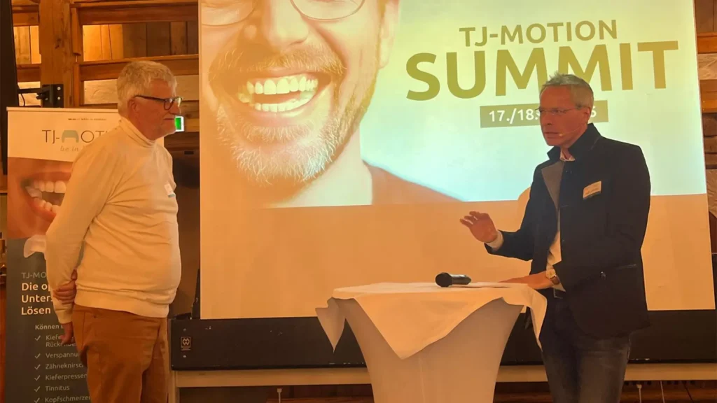 Zwei Referenten sprechen vor einer Leinwand mit der Aufschrift „TJ-MOTION SUMMIT“; vorne steht ein Stehtisch mit Mikrofon, links ein TJ-MOTION-Banner.