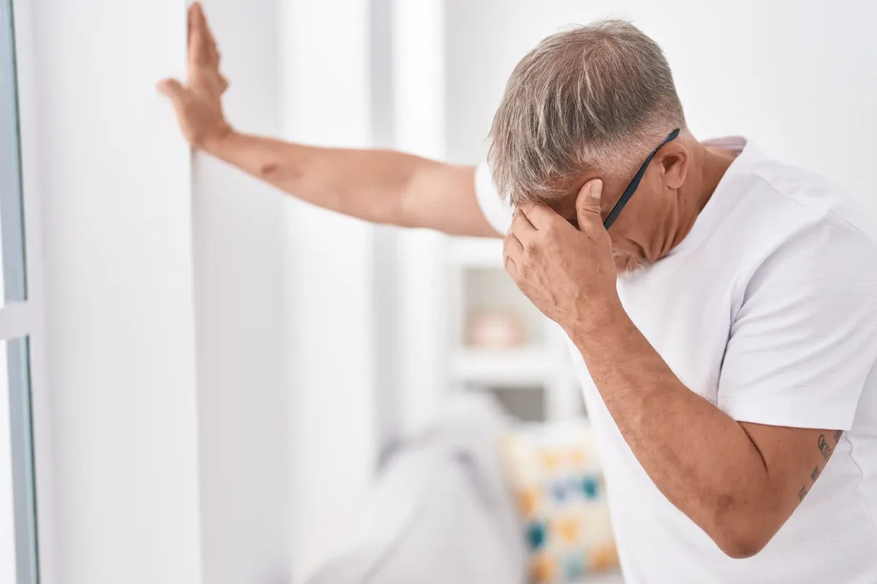 Älterer Mann steht unsicher in einem Raum, stützt sich mit einer Hand an der Wand ab und hält sich mit der anderen den Kopf – Anzeichen von Schwindel oder Gleichgewichtsstörung wobei die vestibuläre Therapie helfen kann.