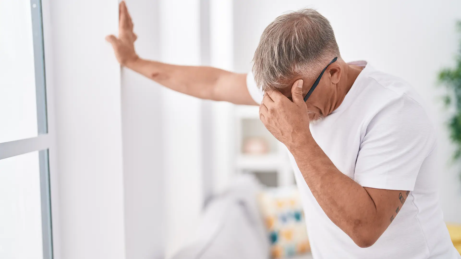 Älterer Mann steht unsicher in einem Raum, stützt sich mit einer Hand an der Wand ab und hält sich mit der anderen den Kopf – Anzeichen von Schwindel oder Gleichgewichtsstörung wobei die vestibuläre Therapie helfen kann.