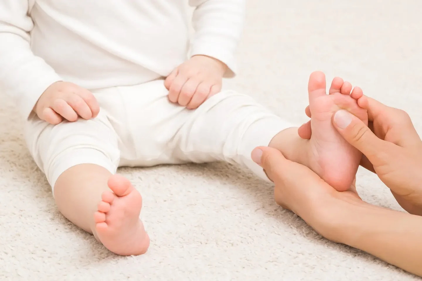 Baby sitzt auf einem Teppich, während eine Person im Rahmen einer Kindertherapie bzw. physiotherapeutischen Behandlung behutsam den Fuß des Kindes hält.