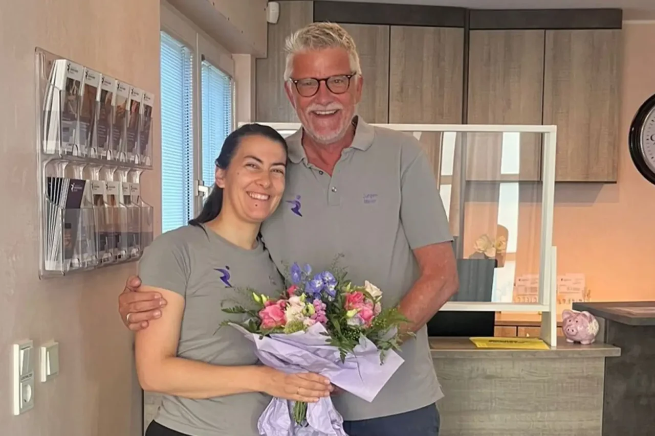 Mitarbeiterin der Physiotherapiepraxis Rösrath steht lächelnd mit Blumenstrauß und einem Kollegen im Empfangsbereich nach erfolgreich abgeschlossener Kinder-Bobath-Ausbildung.