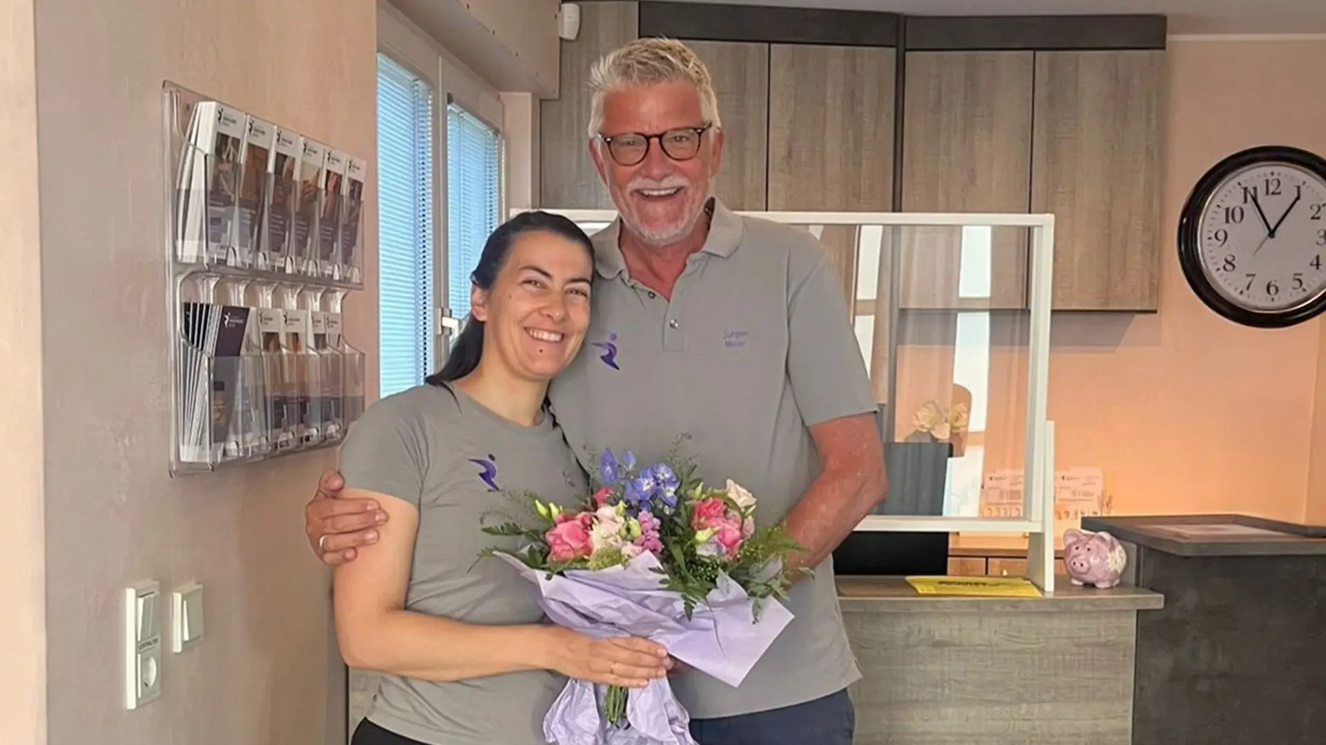 Mitarbeiterin der Physiotherapiepraxis Rösrath steht lächelnd mit Blumenstrauß und einem Kollegen im Empfangsbereich nach erfolgreich abgeschlossener Kinder-Bobath-Ausbildung.