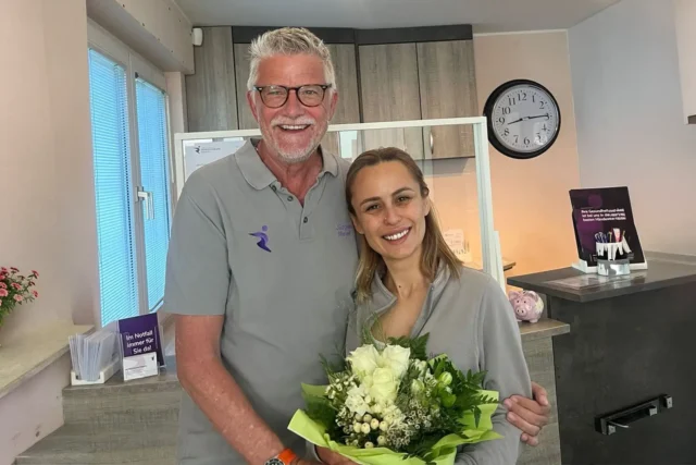 Zwei Mitarbeitende der Physiotherapiepraxis Rösrath stehen lächelnd mit Blumenstrauß im Empfangsbereich nach erfolgreich bestandener Bobath-Weiterbildung.