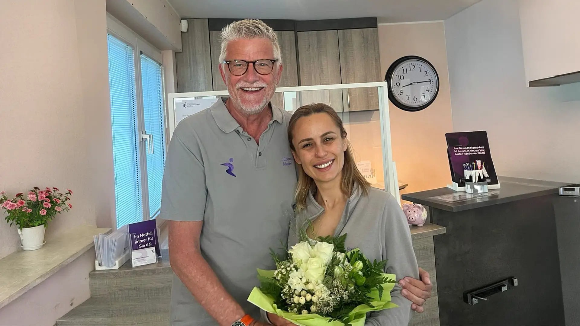 Zwei Mitarbeitende der Physiotherapiepraxis Rösrath stehen lächelnd mit Blumenstrauß im Empfangsbereich nach erfolgreich bestandener Bobath-Weiterbildung.